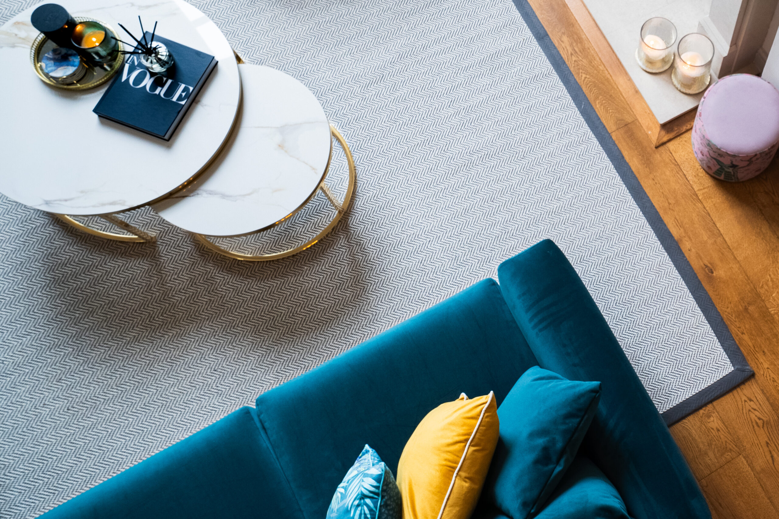 Overhead view of a herringbone rug with a dark border beside a teal sofa and marble coffee tables